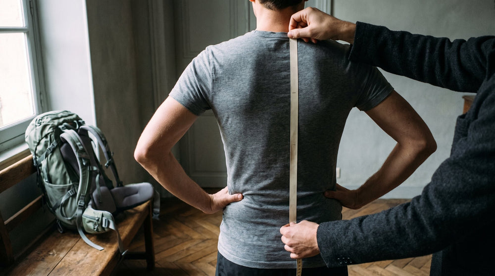 Back view: a helper runs a tape measure from the base of the neck to the hip line, a sage green backpack waiting on a bench
