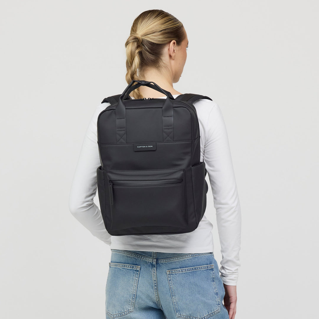 Woman wearing the all black backpack on her back, casual white long-sleeve outfit, structured square silhouette