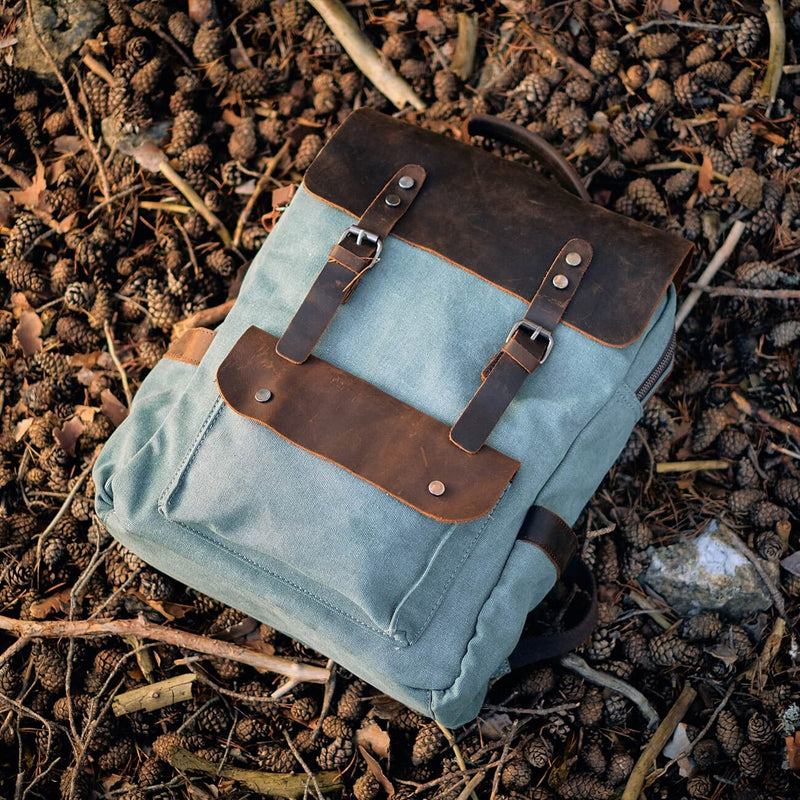 brown leather and light blue canvas backpack laying on forest pine cones ground