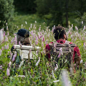two hikers wearing vintage canvas backpacks sat on a flower field at sunset