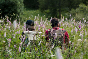 two hikers wearing vintage canvas backpacks sat on a flower field at sunset