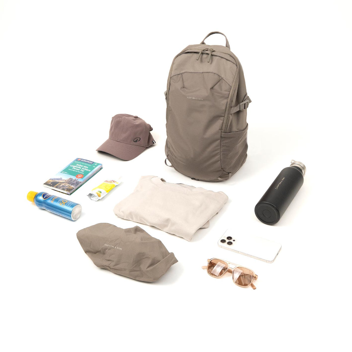 Taupe Åre Daypack Lite with what it carries for a day out: cap, guidebook, sun cream, light top, water bottle, phone, sunglasses and its rain cover