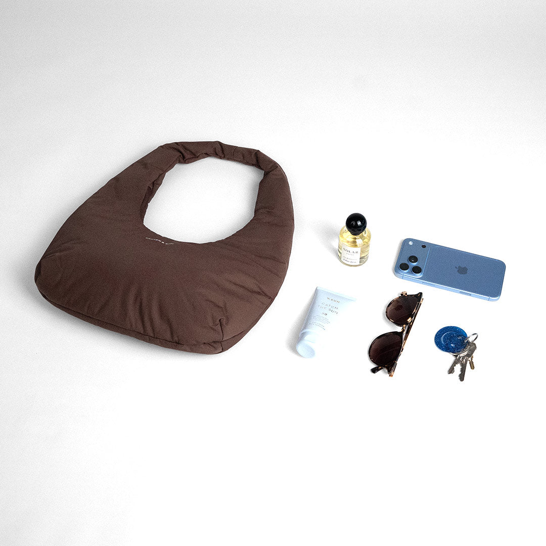 Chocolate brown Skara Cloud Shoulder with a phone, keys, sunglasses, a perfume and a tube of sun cream laid out beside it