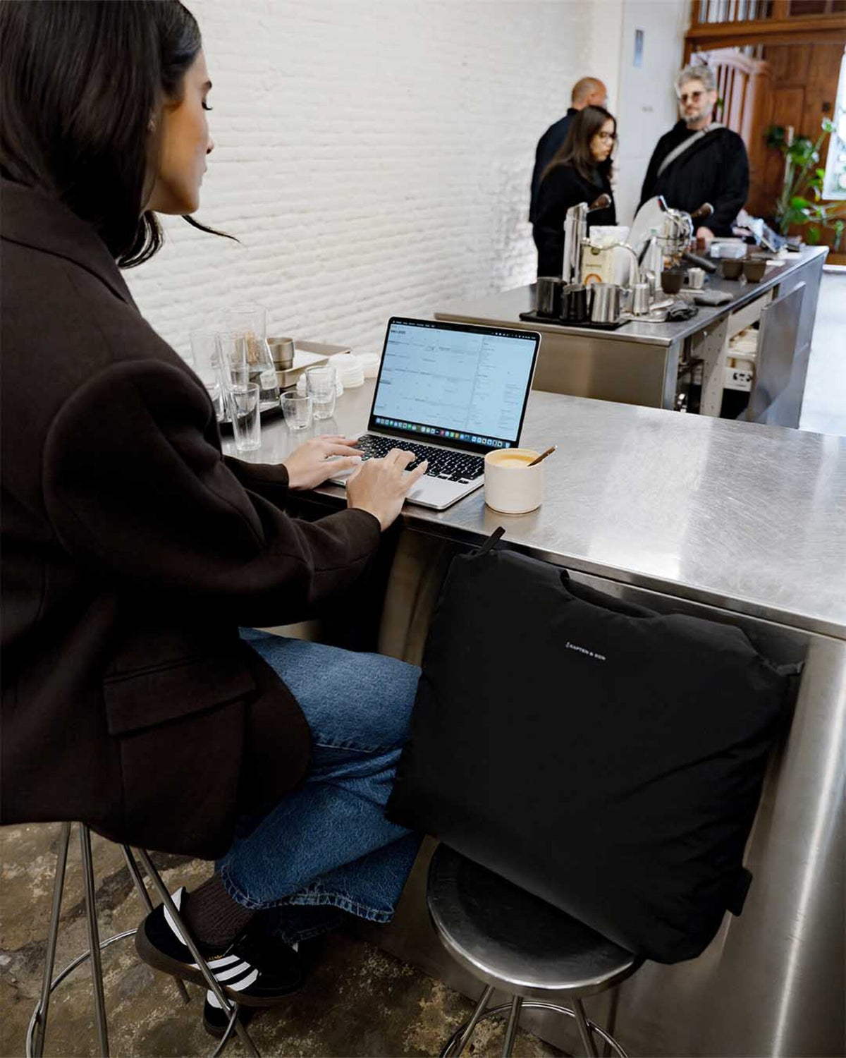Woman working on a laptop at a café counter, the black Skara Cloud Tote on the stool beside her