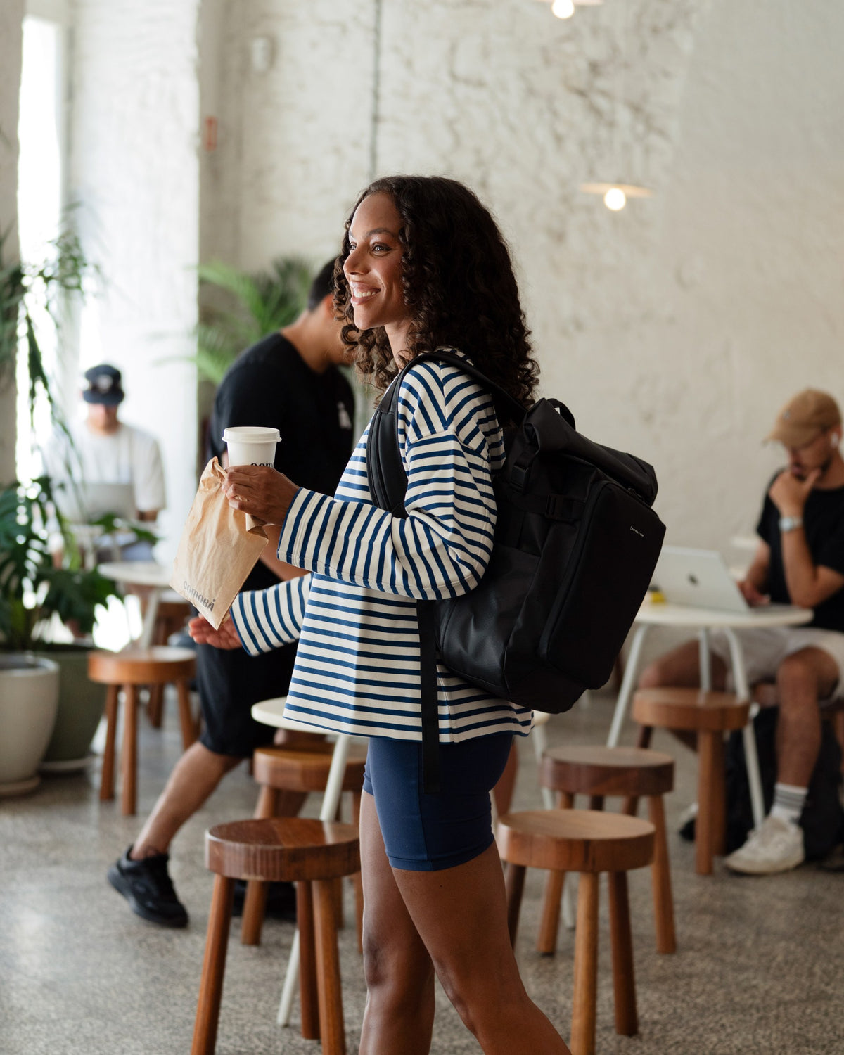 Woman in striped sweater carrying the Lisbon Small black with a takeaway coffee in a café