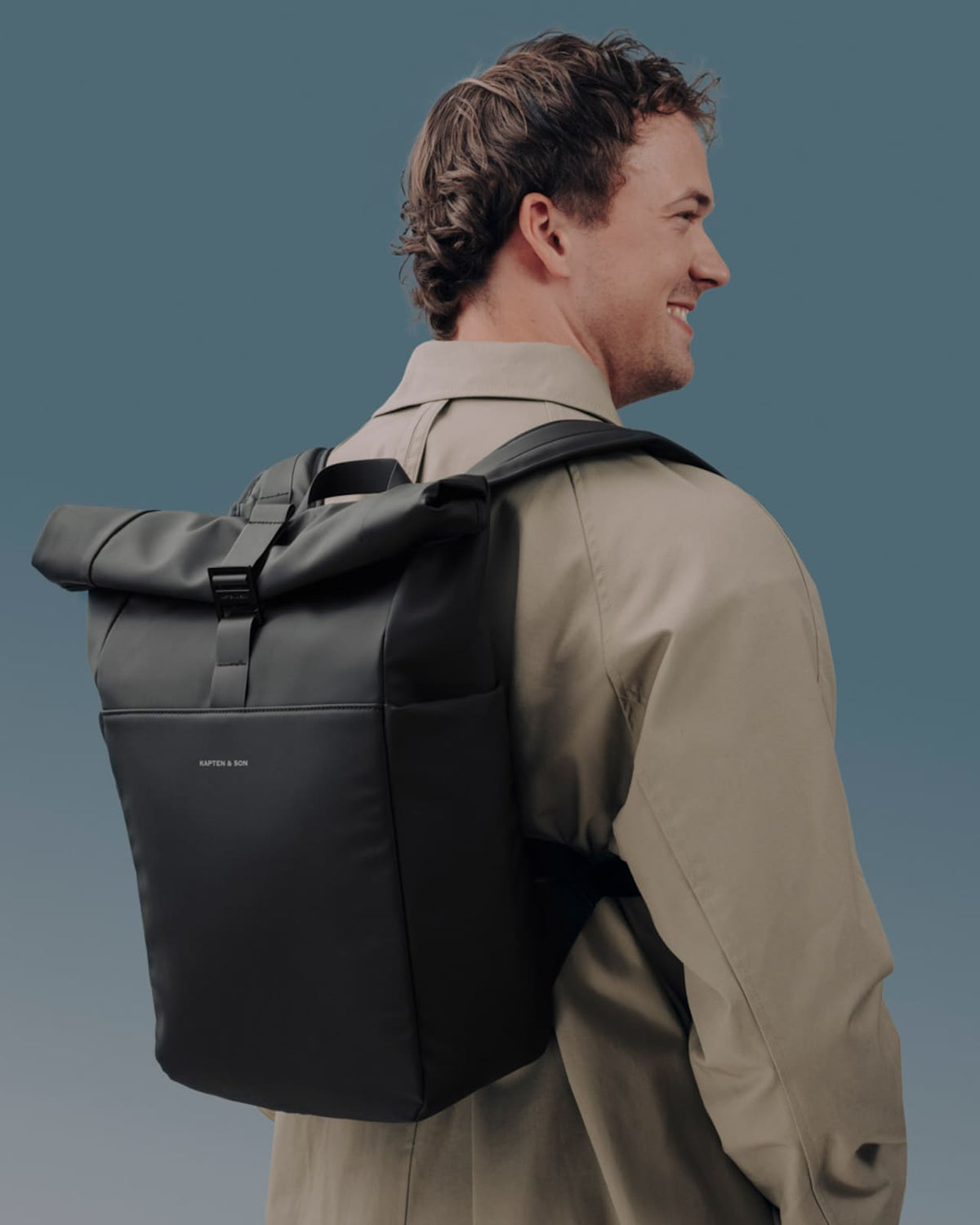 Man wearing the all black rolltop backpack on one shoulder, profile view, neutral trench outfit