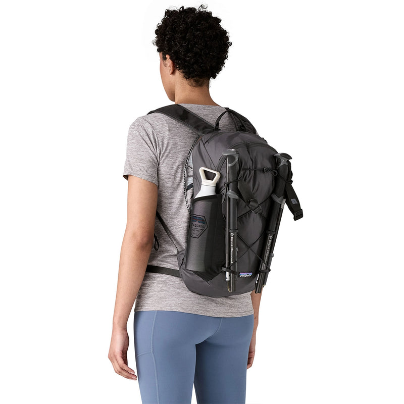 Woman carrying a loaded black trail pack with trekking poles and bottle, ready for outdoor adventure