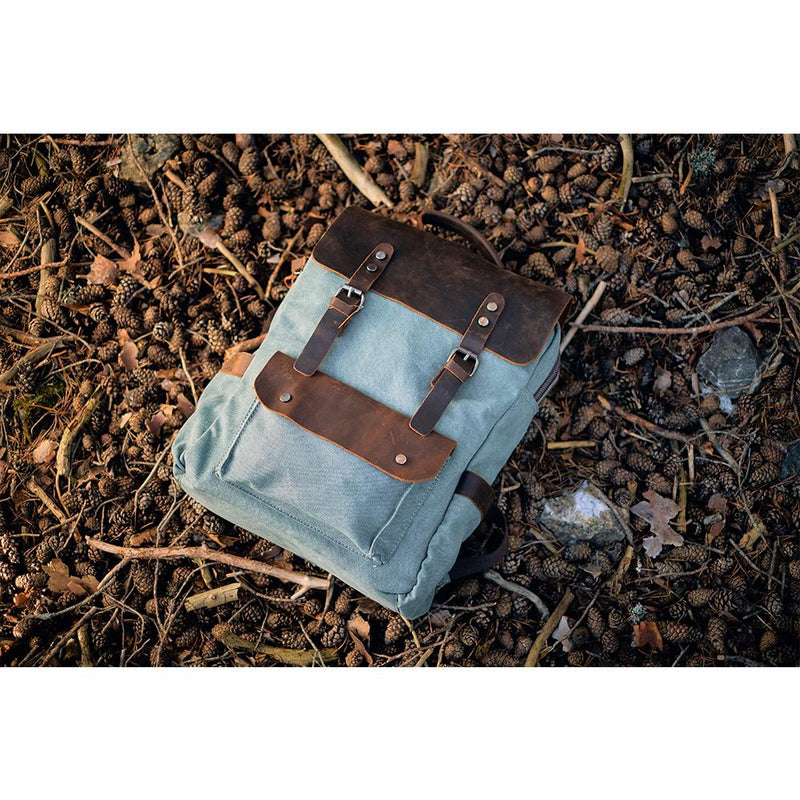 brown leather and light blue canvas backpack laying on forest pine cones ground